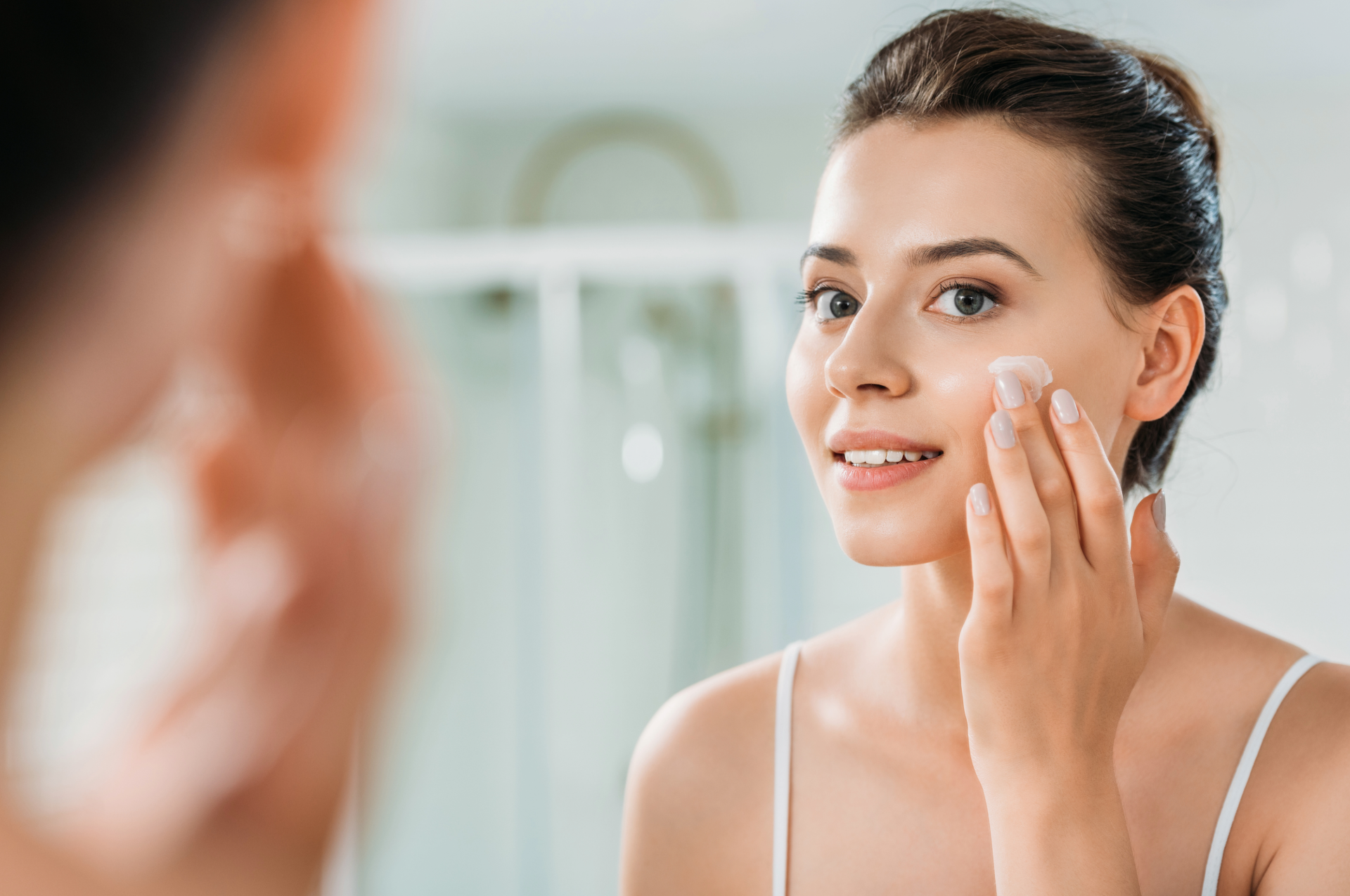 Woman applying cream to her face in front of a mirror, bathroom setting, smiling.
