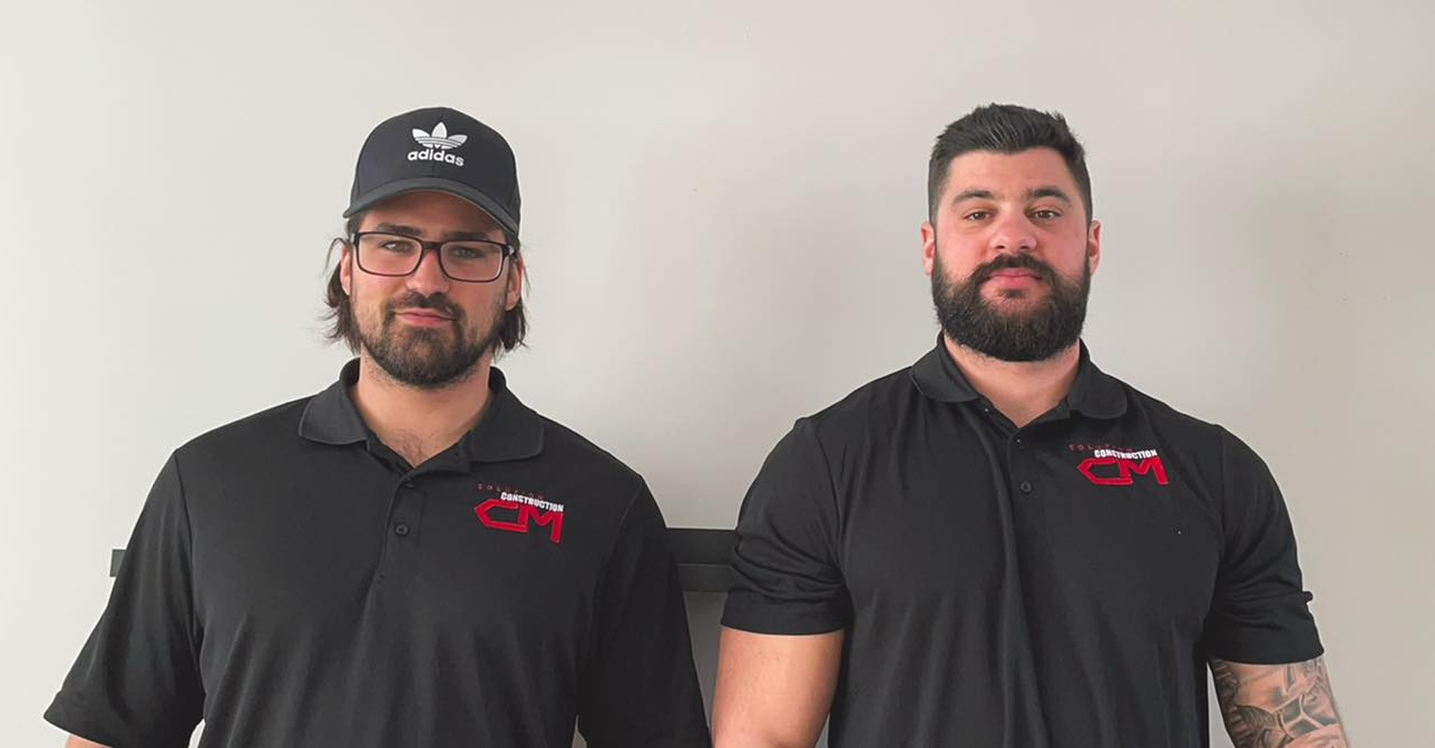 Deux hommes en polos noirs se tiennent devant un mur clair et uni, l'un d'eux porte une casquette noire.
