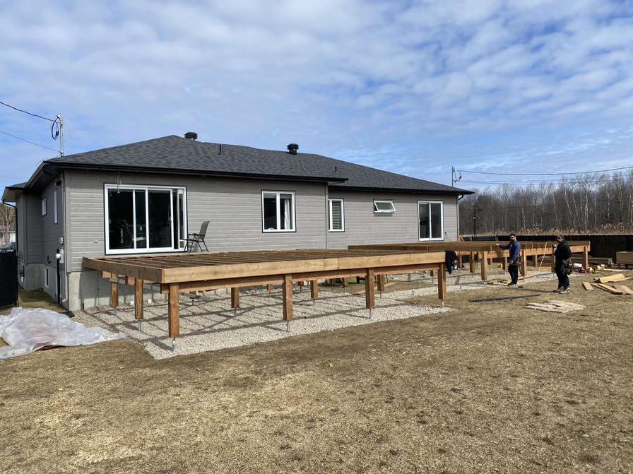 Construction d'une terrasse en bois sur gravier à côté d'une maison grise ; les gens travaillent à l'extérieur.