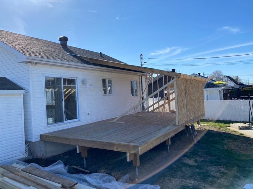 Construction d'une terrasse en bois attenante à une maison blanche avec une extension de toit partiellement construite sous un ciel bleu.