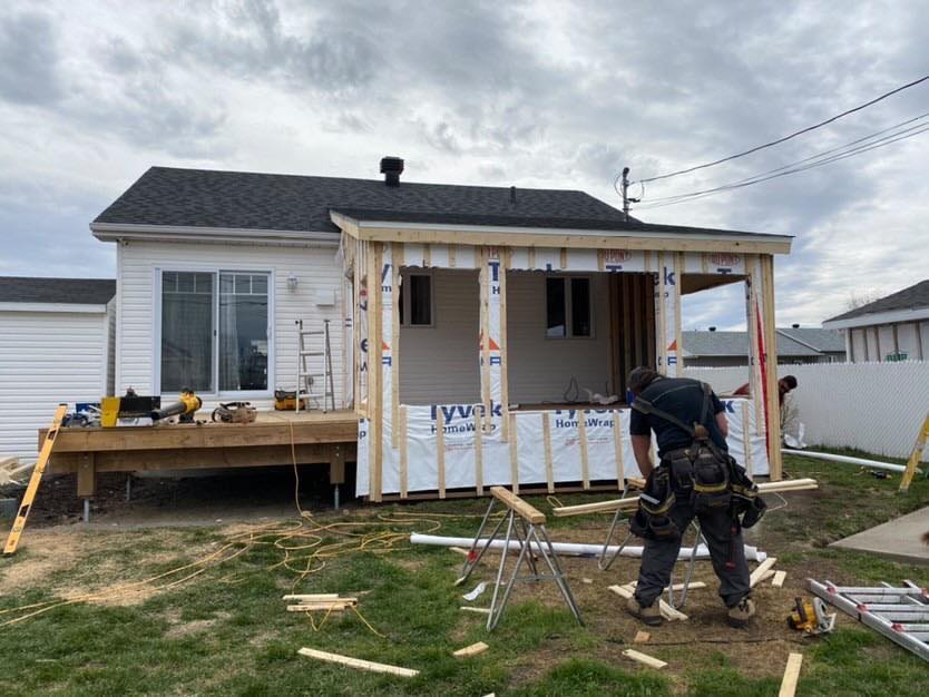 Un ouvrier du bâtiment construit un porche fermé pour une maison blanche par une journée nuageuse.