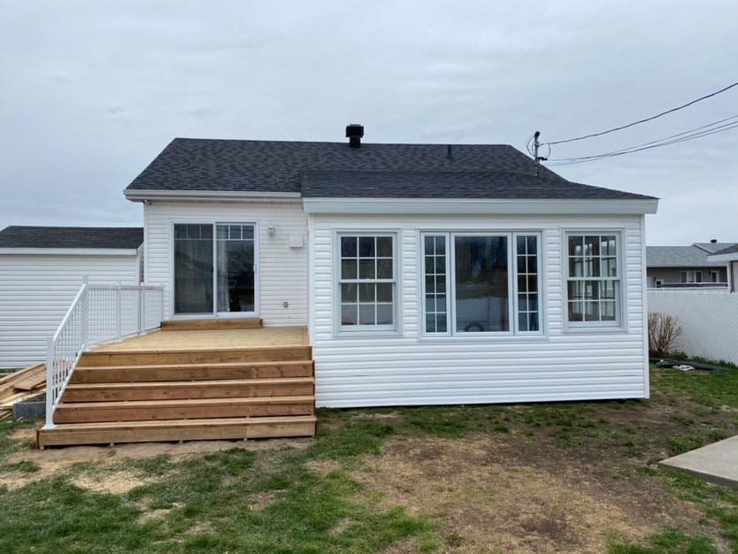 Maison blanche avec terrasse en bois et grandes fenêtres, posée sur une herbe verte sous un ciel nuageux.