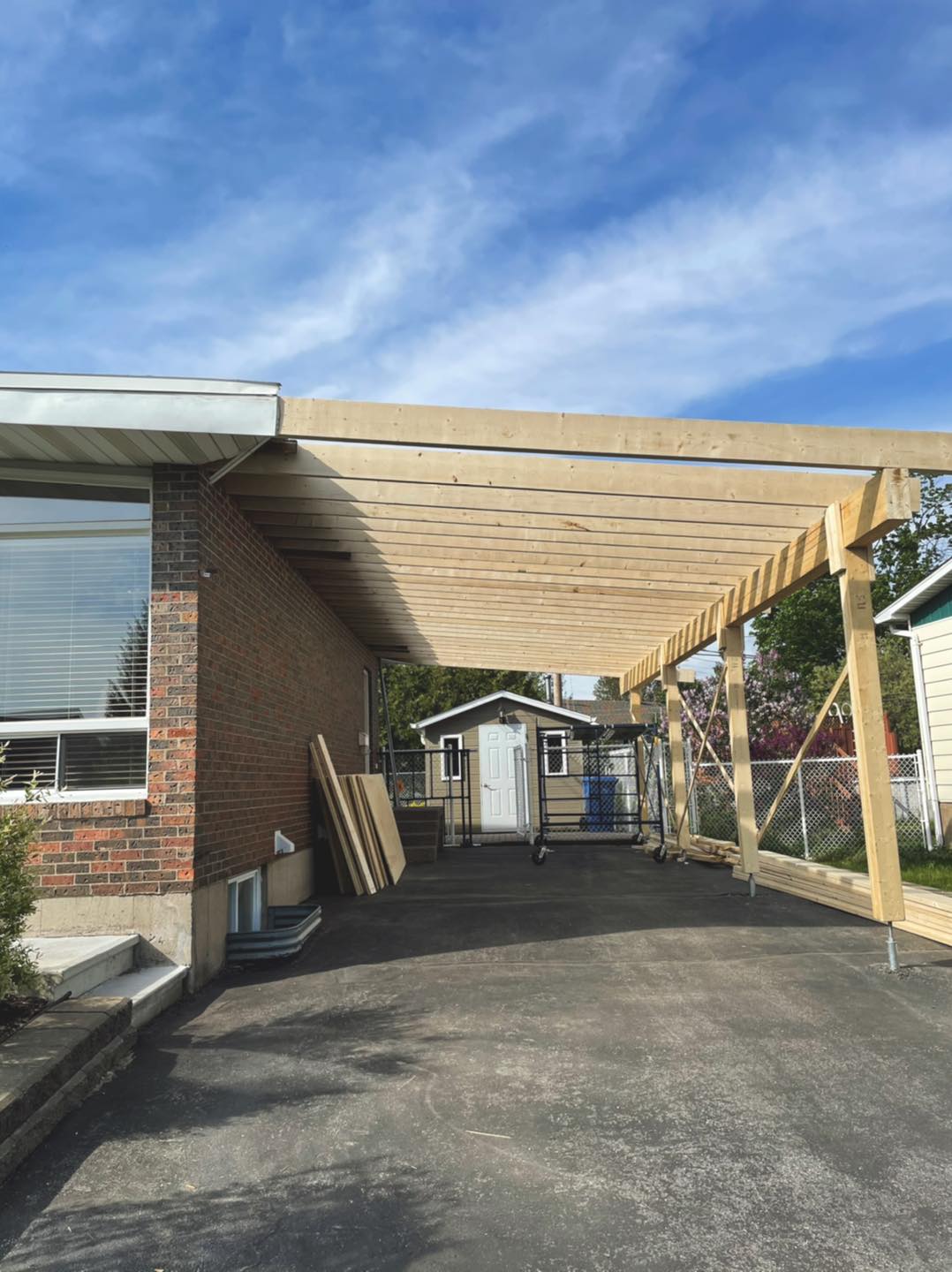 Abri d'auto en construction à côté d'une maison en briques, avec un ciel clair en arrière-plan.
