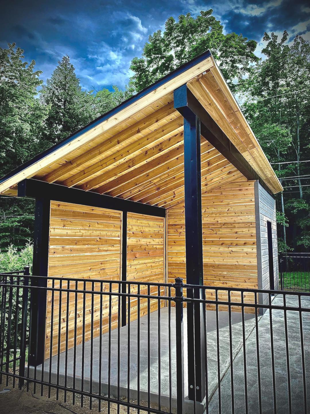 Un abri en bois avec supports et toit noirs, accessible par une balustrade en métal noir.