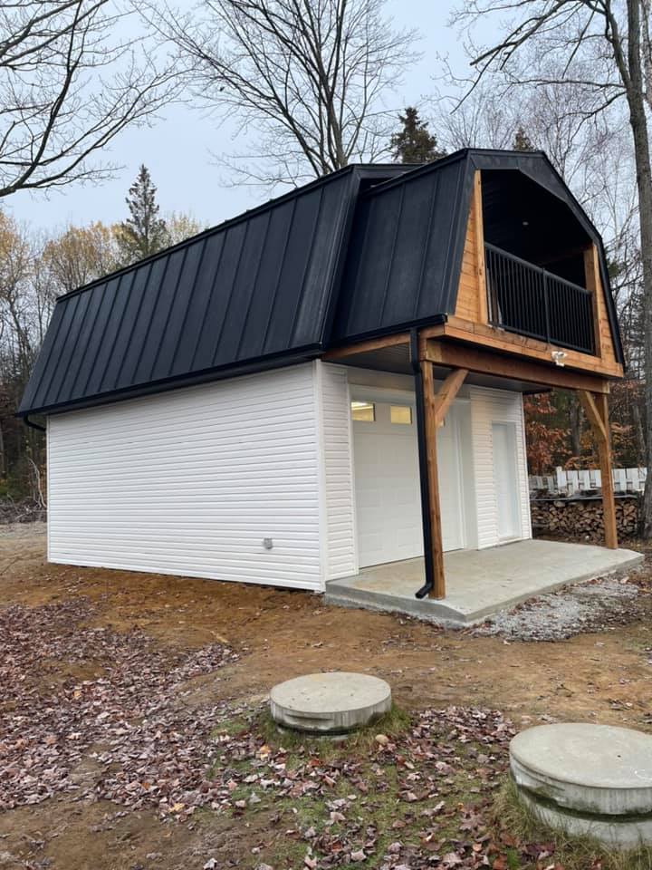 Un garage indépendant blanc pour deux voitures, avec un toit à deux pans brisés, une toiture en métal noir et un balcon à l'étage, dans un cadre boisé.