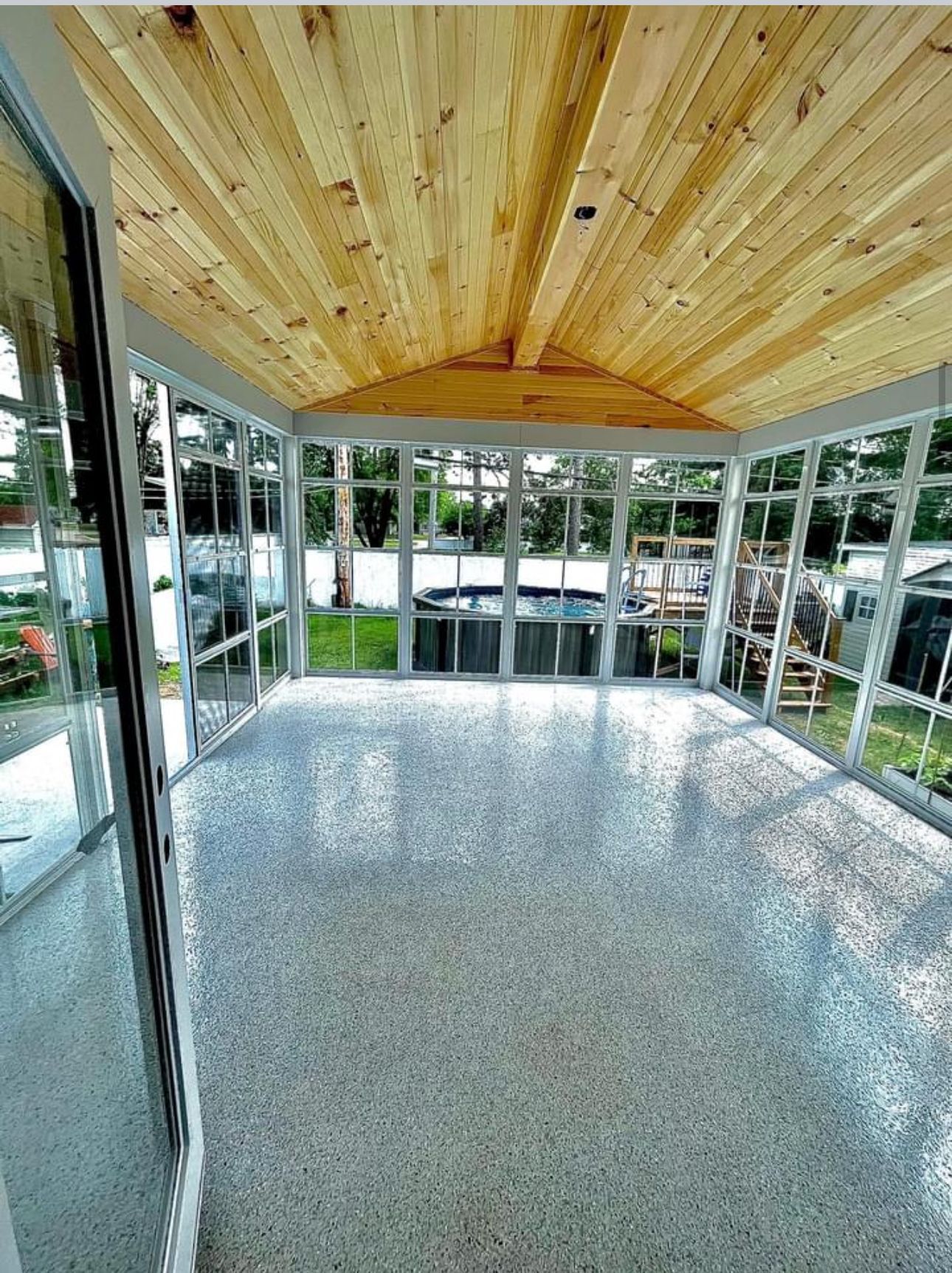 Une véranda avec un plafond lambrissé, des murs en verre et un sol en époxy clair moucheté donnant sur une piscine dans le jardin.