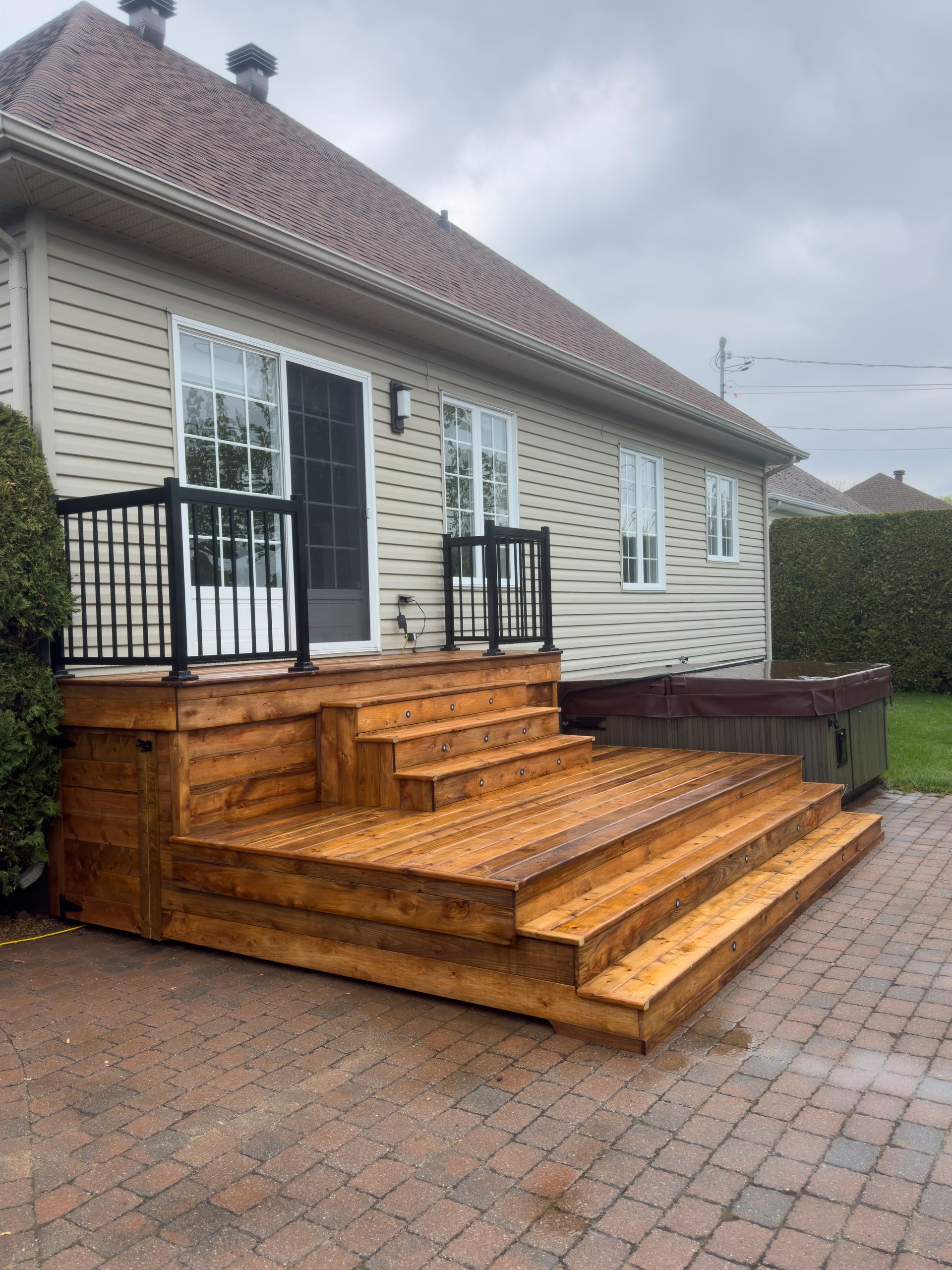 Une terrasse en bois avec des marches menant à la porte d'entrée de la maison, située à côté d'un jacuzzi sur une terrasse pavée.