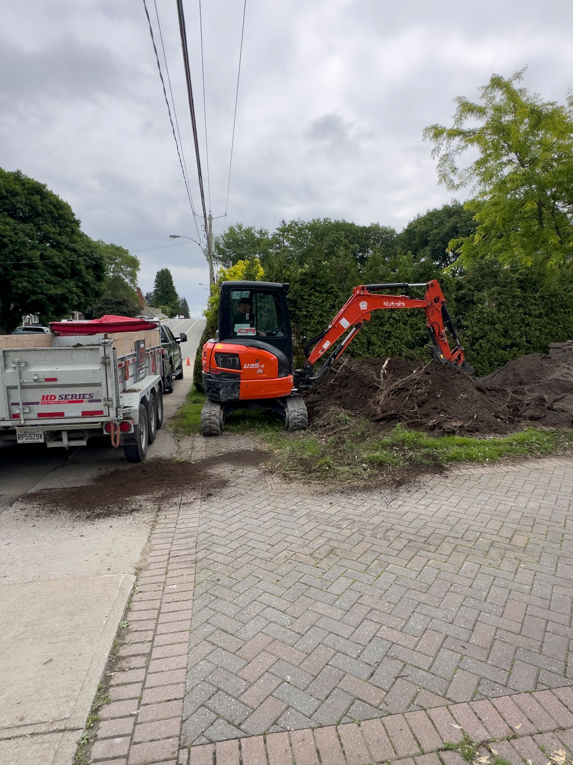 Une pelleteuse orange creuse la terre à côté d'une route pavée et d'une remorque. Des lignes électriques et de la végétation sont visibles.
