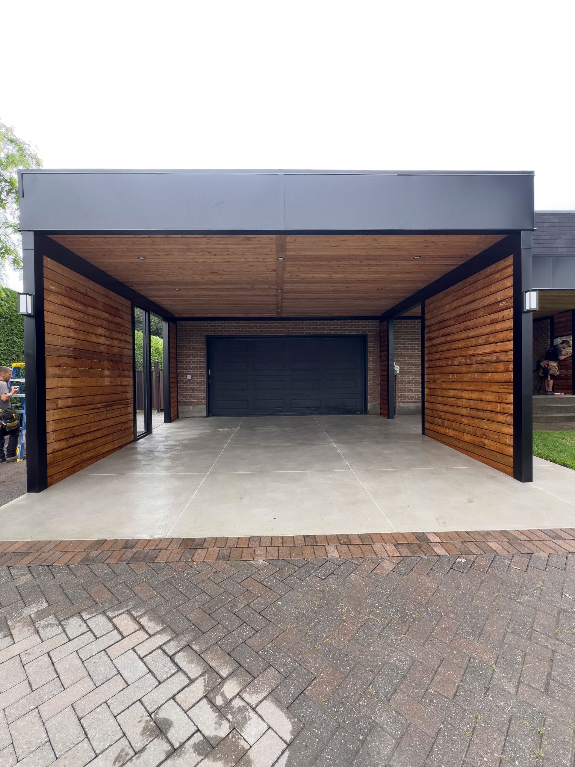 Abri d'auto avec moulures noires, panneaux de bois et porte de garage grise. Sol en béton, allée en briques.