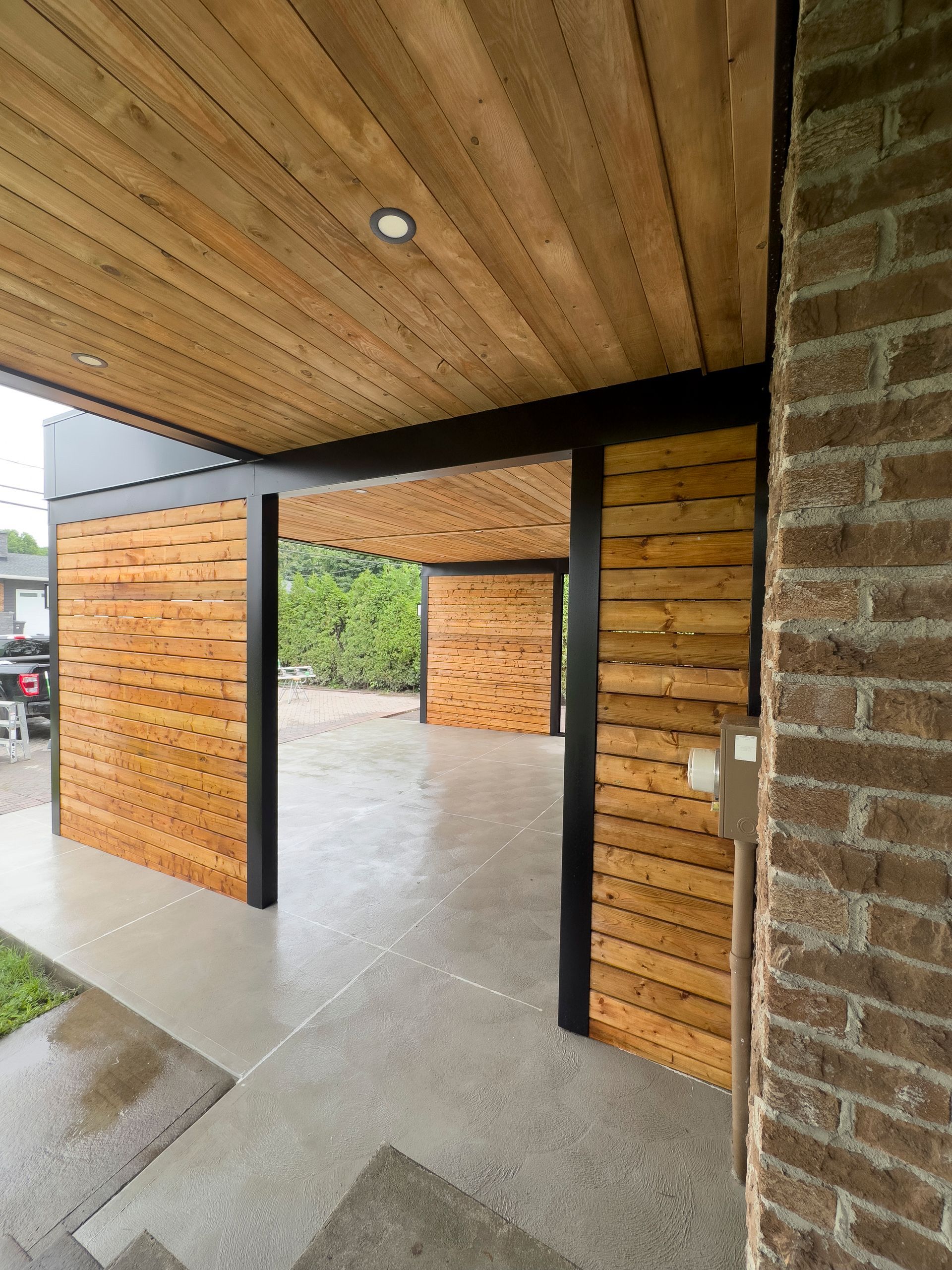 Allée couverte à ciel ouvert avec bardage et plafond en bois, mur en briques et sol en béton.