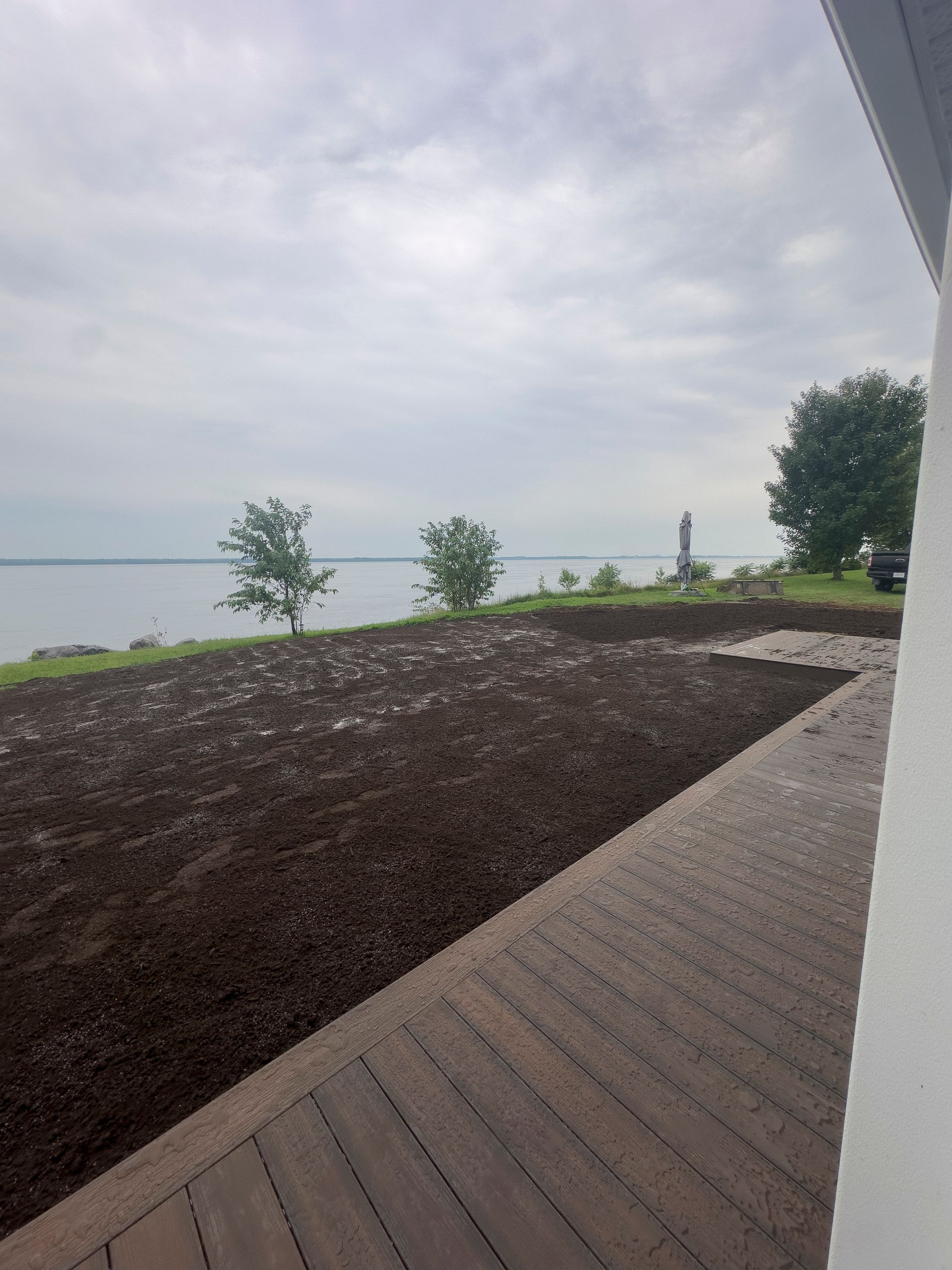 Une parcelle de terre brune jouxte une terrasse en bois donnant sur un lac et un ciel nuageux. Trois petits arbres bordent la rive.