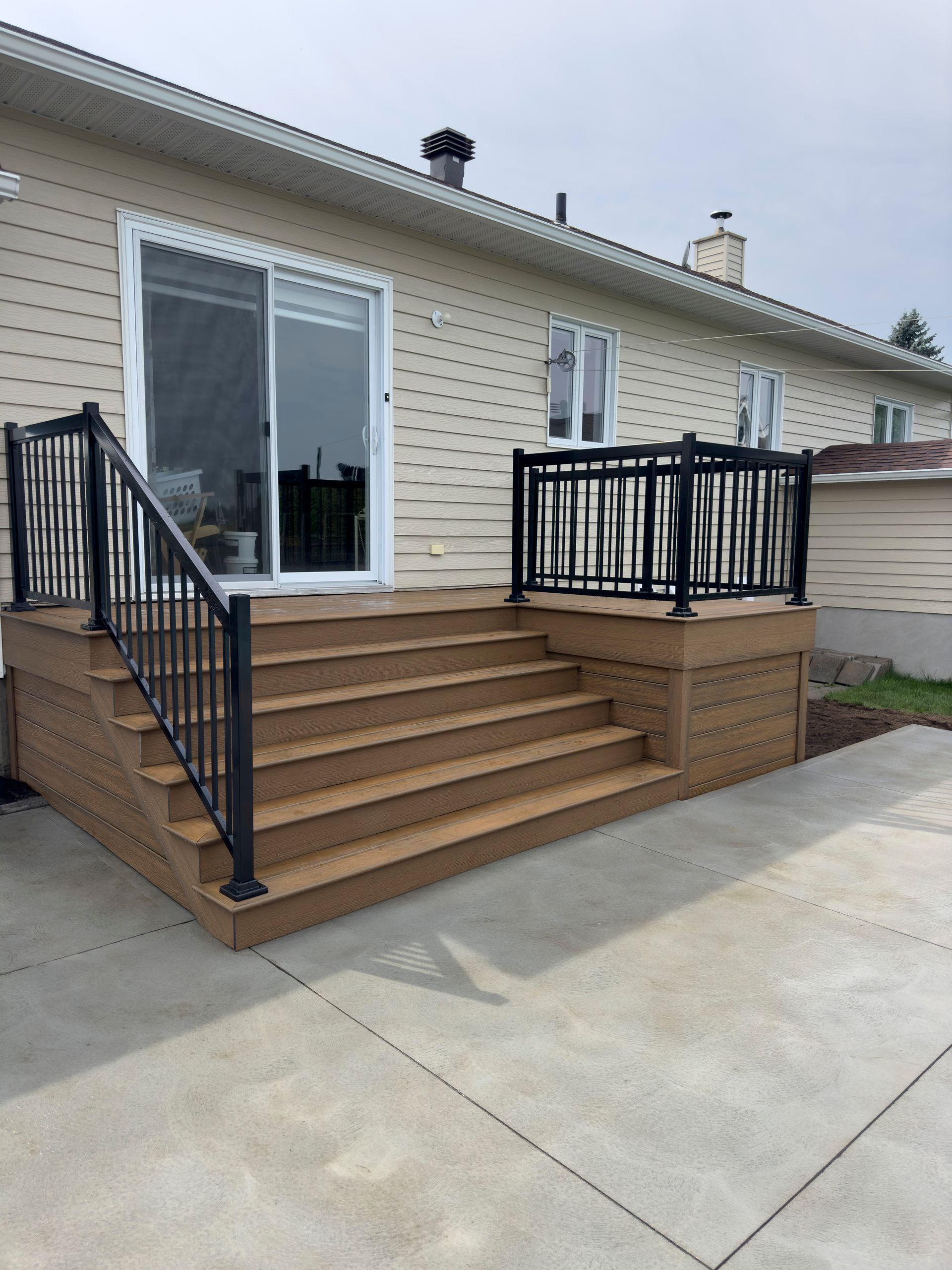 Terrasse en bois marron avec balustrade noire et marches menant à une porte coulissante en verre.