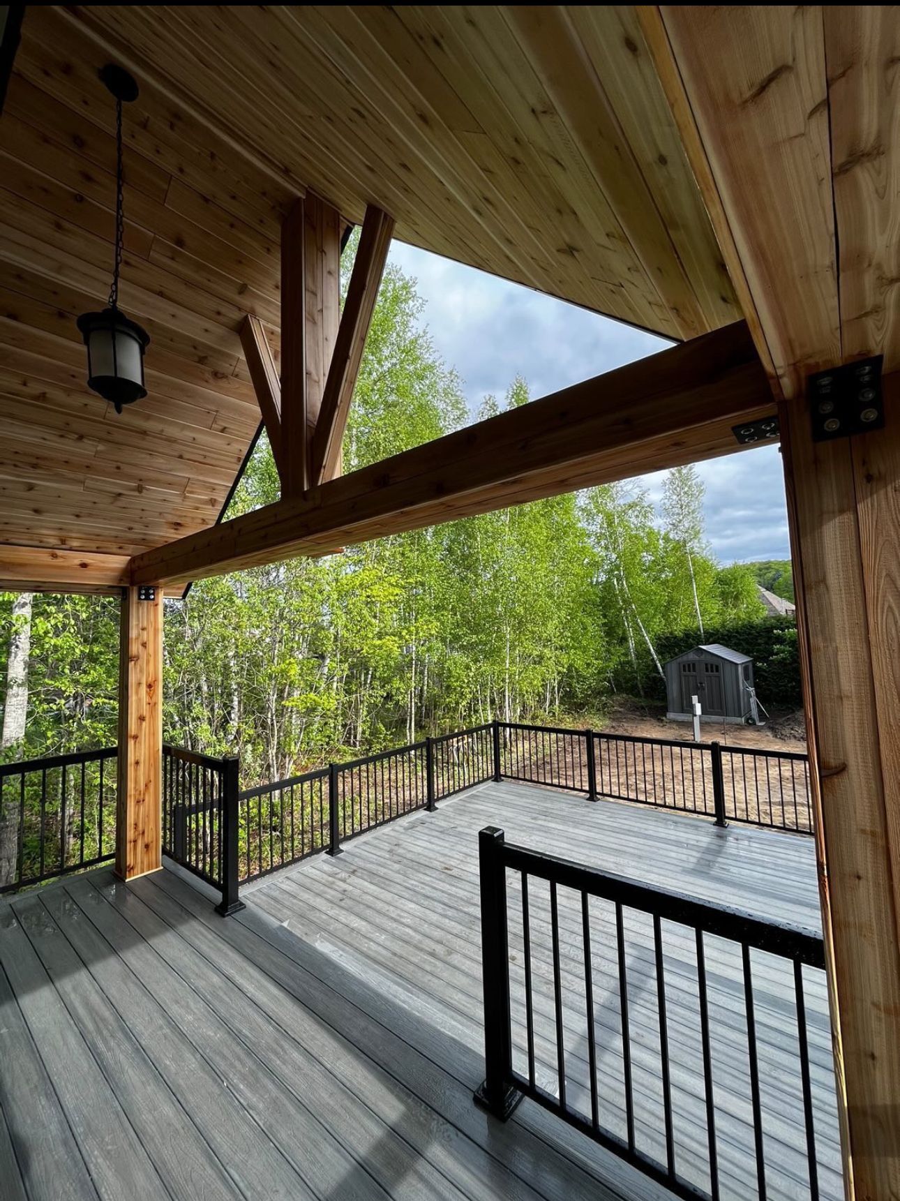 Terrasse en bois avec garde-corps noir, surplombant une forêt. Des poutres en bois soutiennent le toit et une lanterne suspendue.