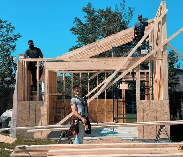 Trois ouvriers du bâtiment construisent une structure en bois à l'extérieur, sous un ciel bleu dégagé.