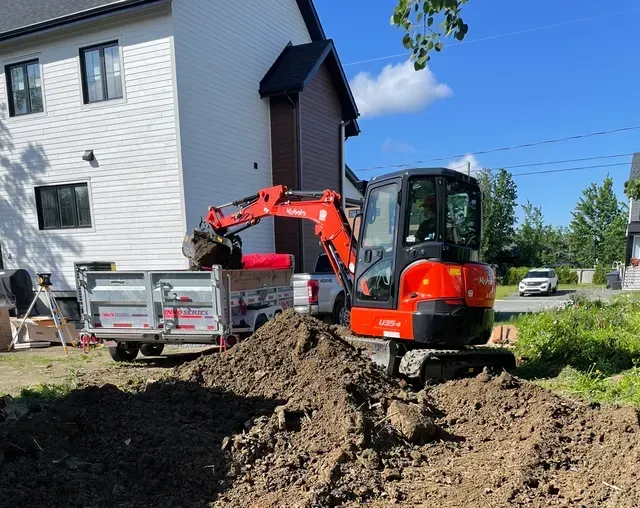 Une pelleteuse orange charge de la terre dans une remorque devant un bâtiment blanc par une journée ensoleillée.