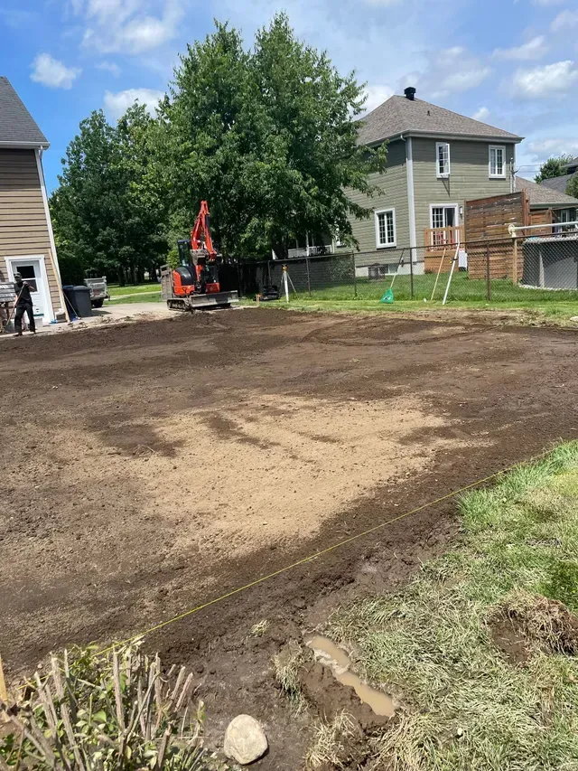 Une petite pelleteuse est en train de creuser un jardin ; de la terre est répandue. Une maison et des arbres se dressent à l’arrière-plan.