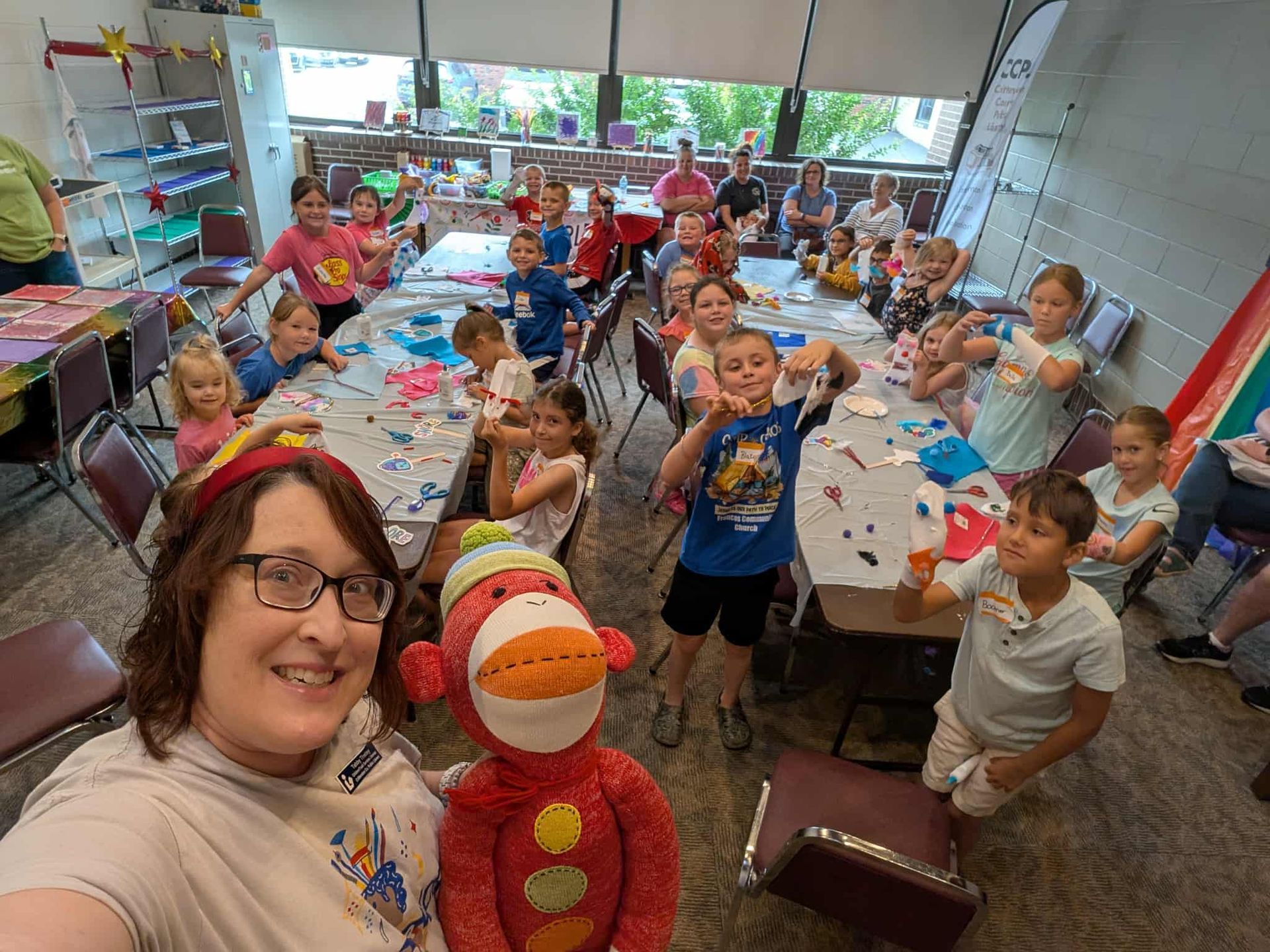Woman and stuffed animal smiling with a group of children at a craft table. Bright, indoor setting.