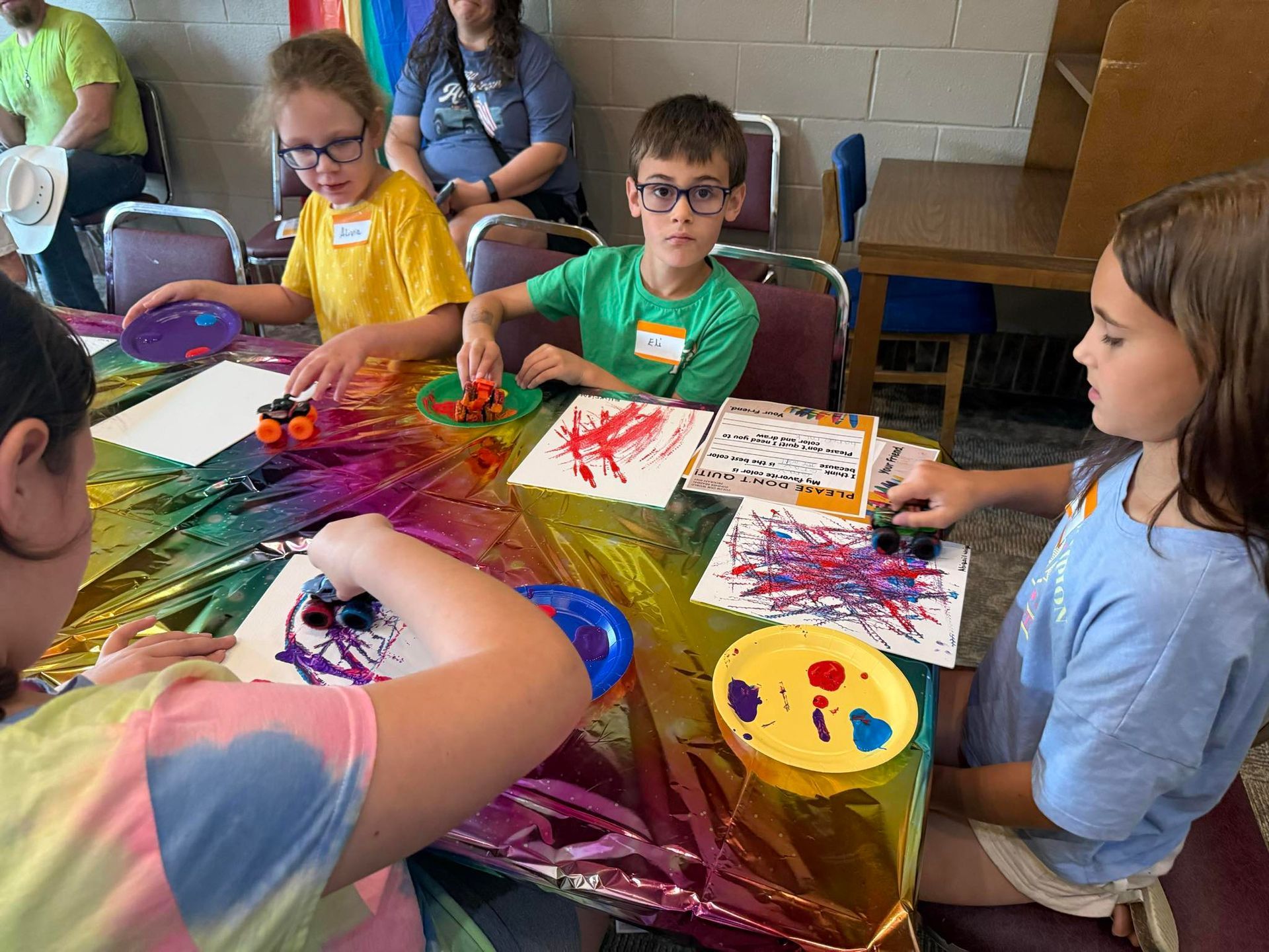 Children painting with toy cars at a table covered in colorful plastic.