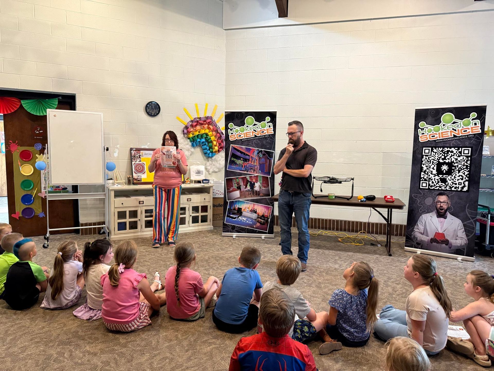 A woman and a man presenting to a group of children seated on the floor in a room.