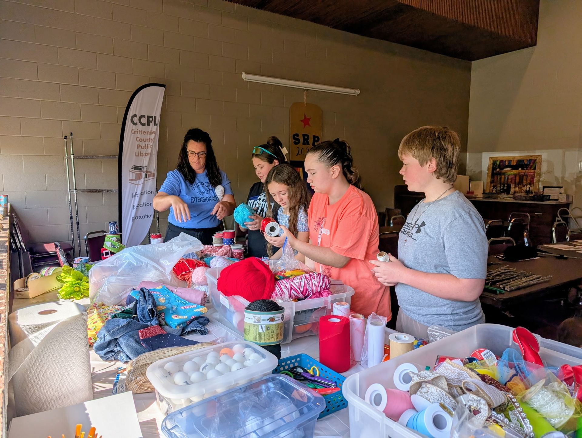 People crafting at a table with yarn, thread, and supplies in an outdoor setting.