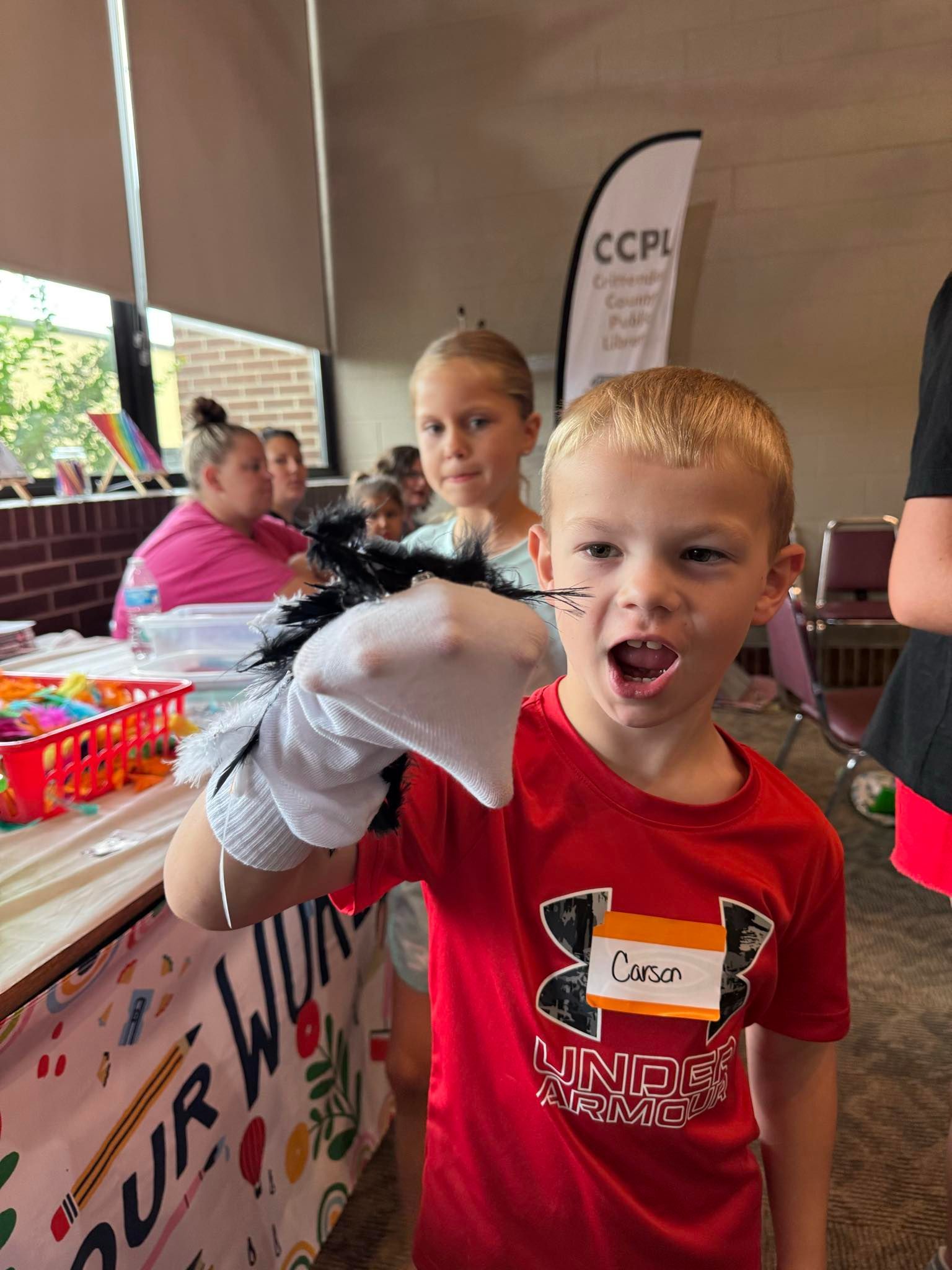 Boy in red shirt with sock puppet, mouth open, indoors at an event, with children and a table.