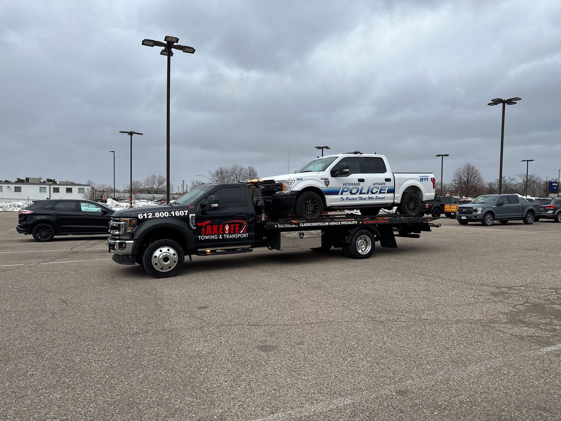 Tow truck carrying a police pickup truck on a gravel lot under a cloudy sky.