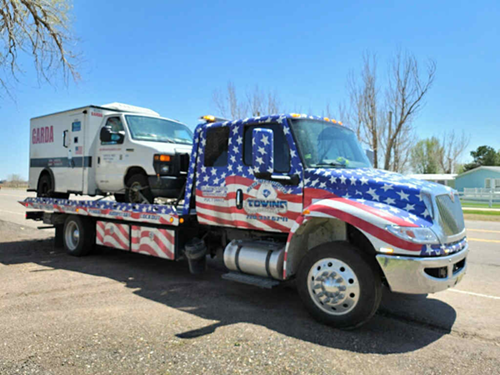 Tow truck with American flag design carrying a Garda armored truck on a sunny road.