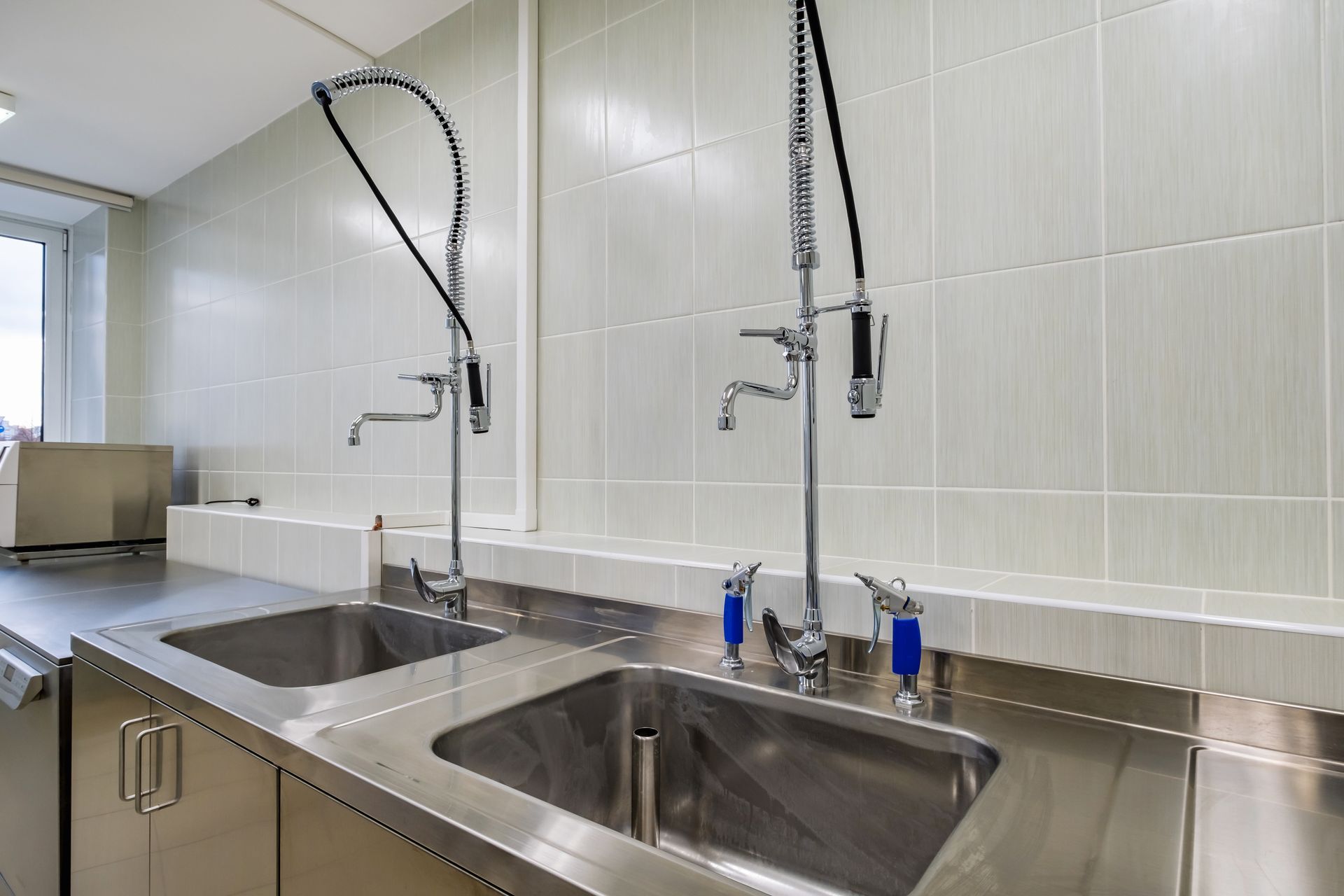 Stainless steel commercial sink area with two high-arc sprayers and tiled wall in a clean kitchen.
