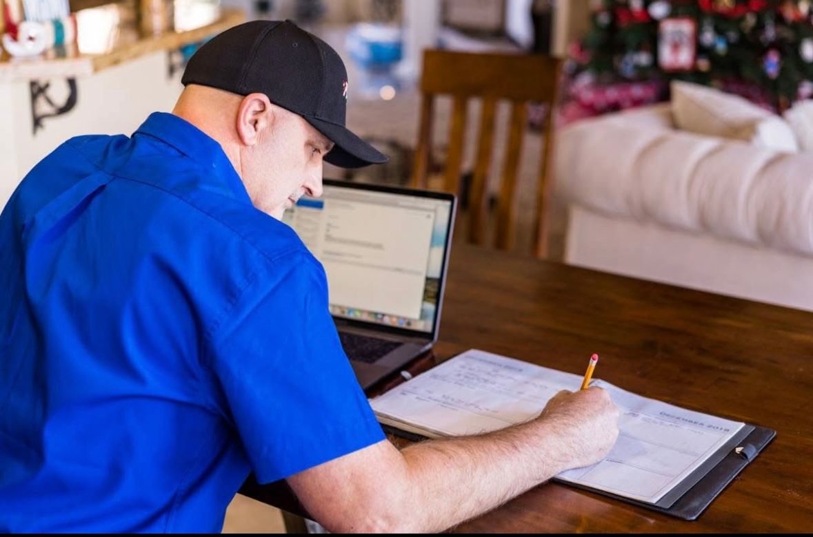Person in a blue shirt and black cap working at a laptop with papers on a table