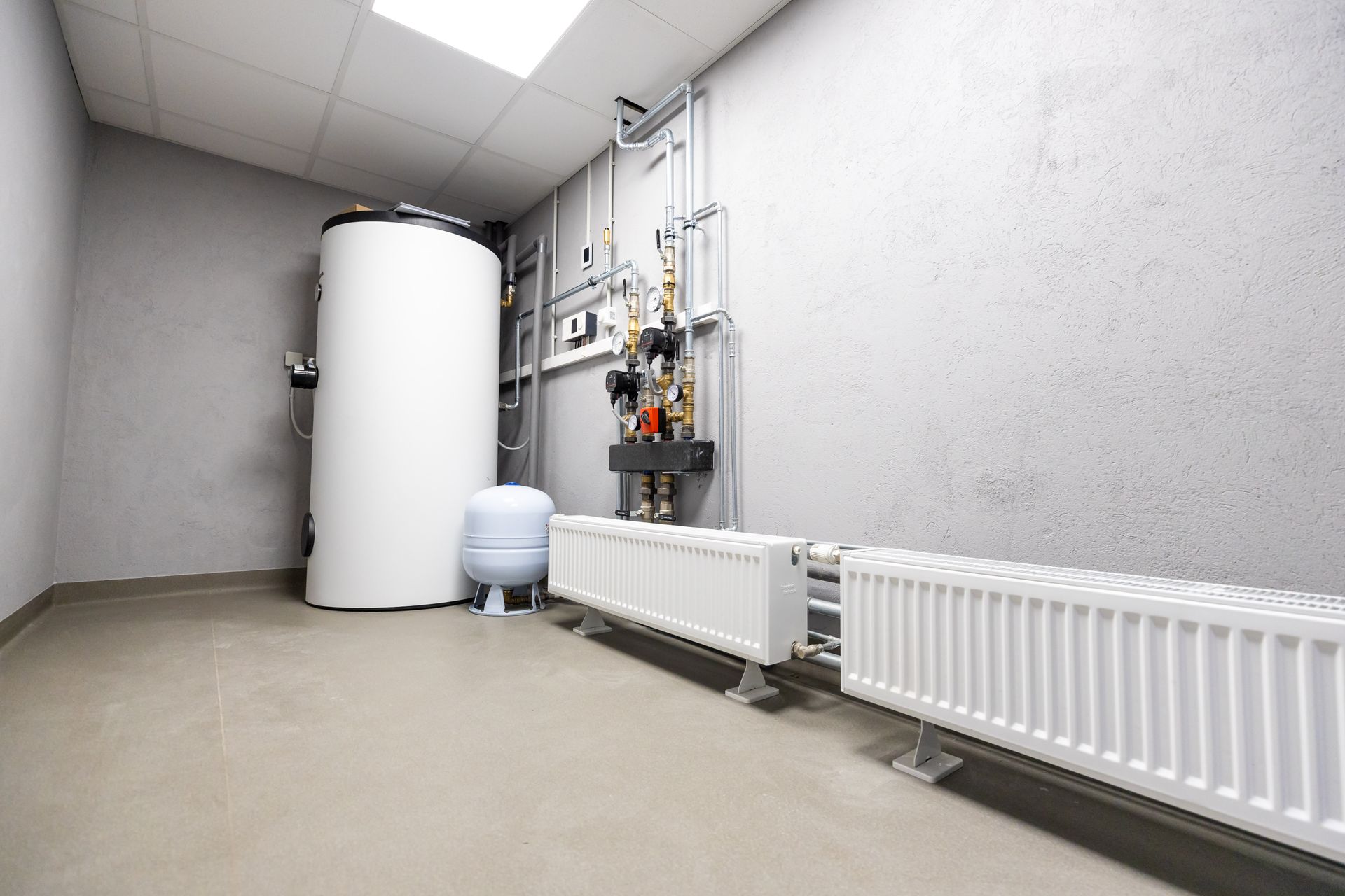 Utility room with white tank, pipes, and radiators along a gray wall