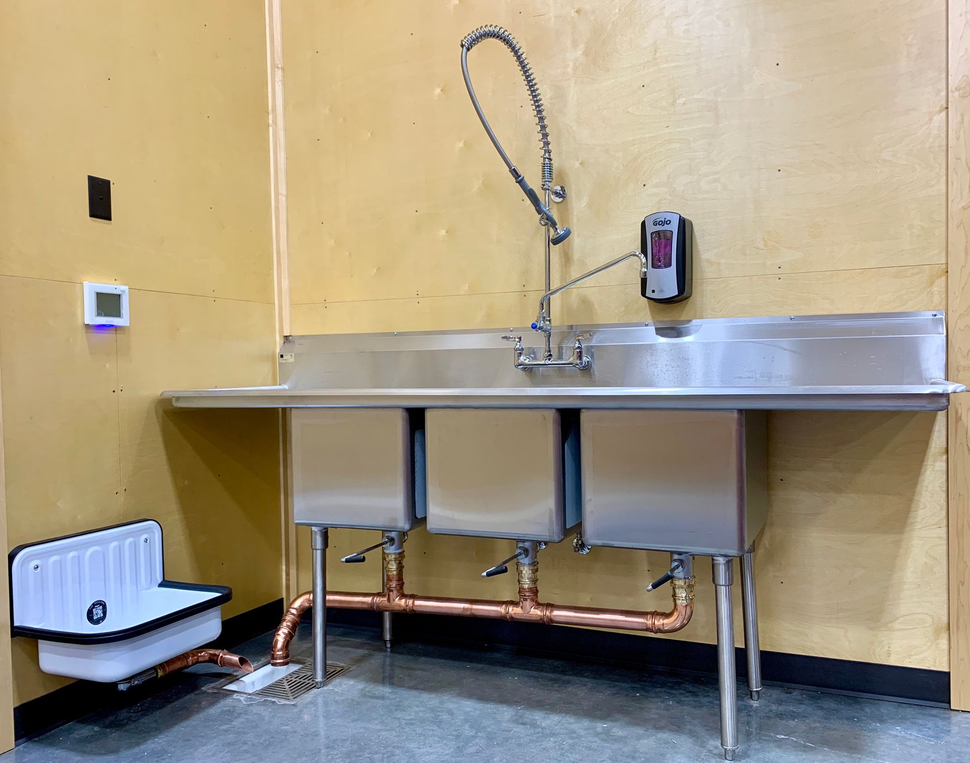 Stainless steel utility sink with wall-mounted sprayer in a yellow room, with a soap dispenser and floor drain.