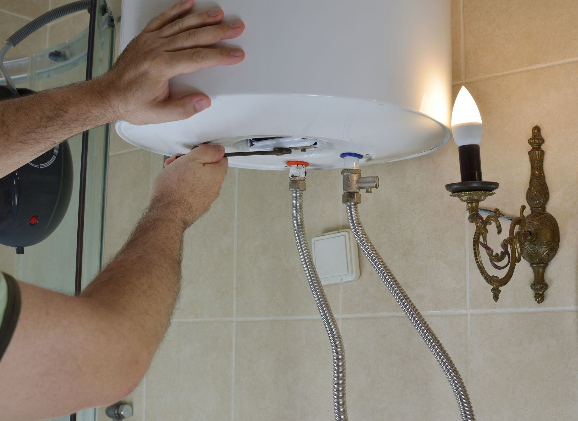 Hands adjusting a white wall-mounted fixture with braided hoses beside a lit wall sconce.