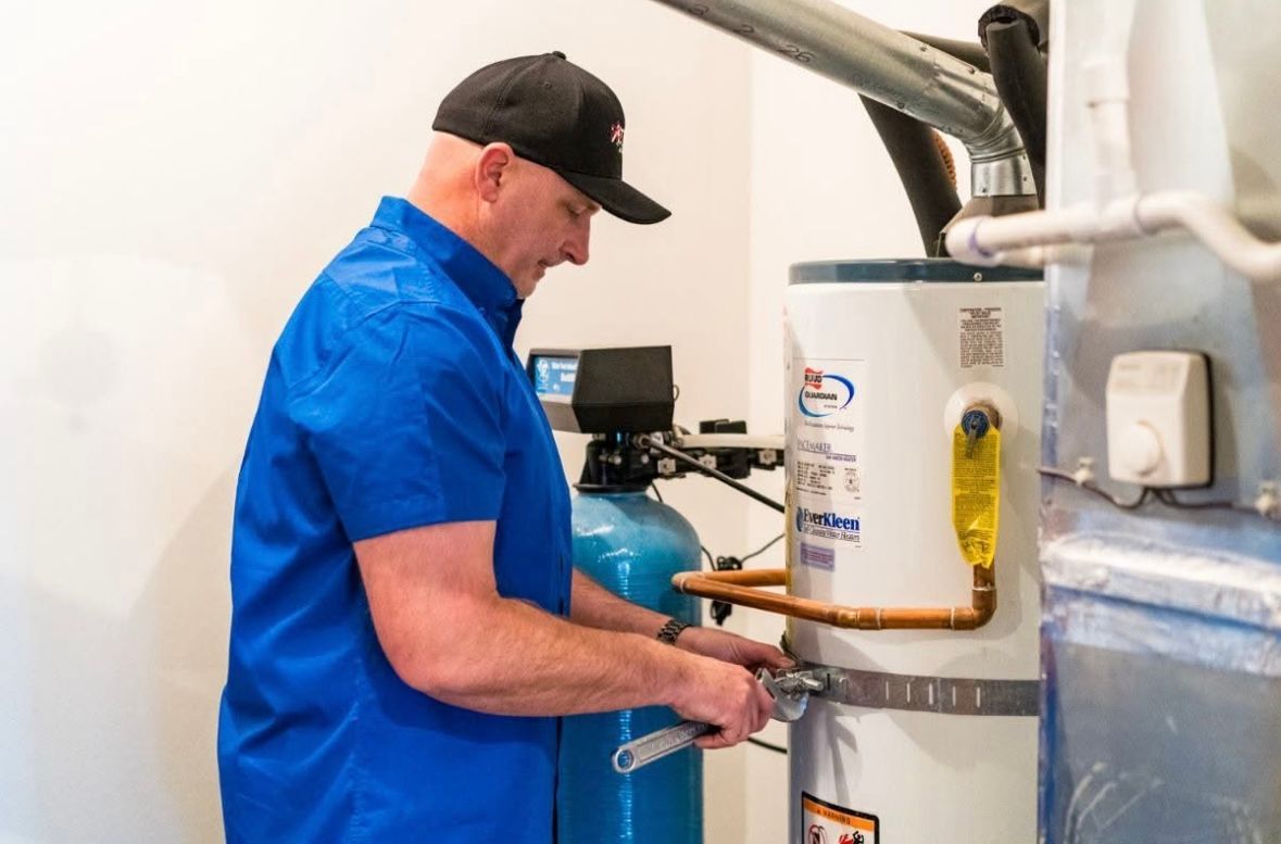 Technician in blue shirt works on a white water heater beside a blue tank in a utility room