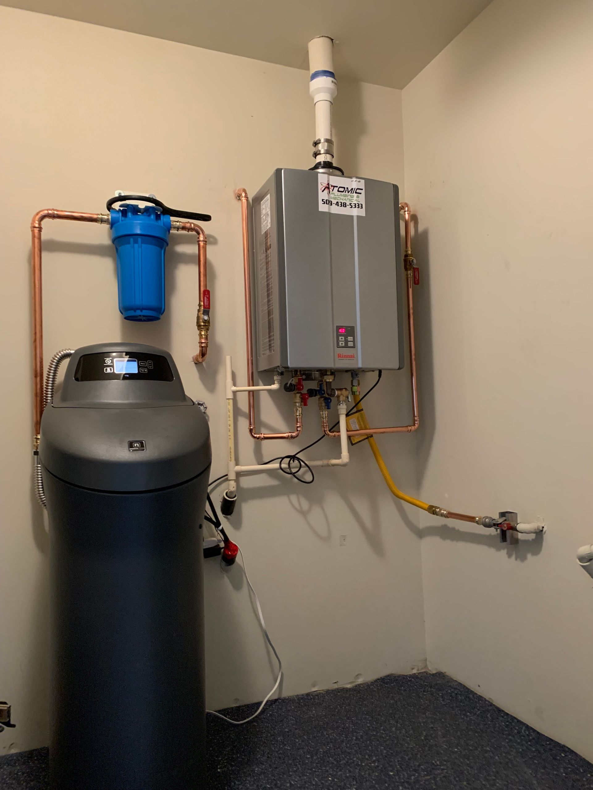 Water softener and tank beside a wall-mounted gas water heater in a utility room.