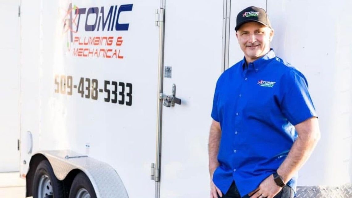 A man in a blue shirt is standing in front of an atomic plumbing and mechanical trailer