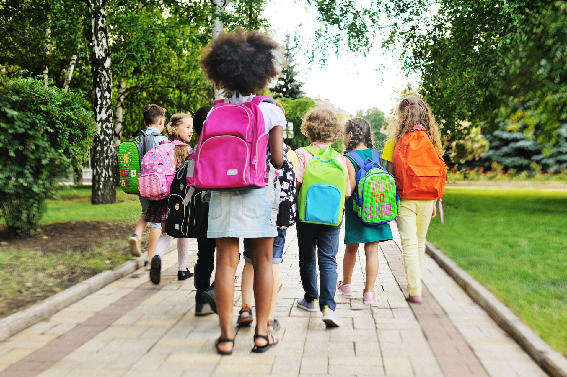 A group of children with backpacks are walking down a sidewalk. A group of children with backpacks are walking down a sidewalk.