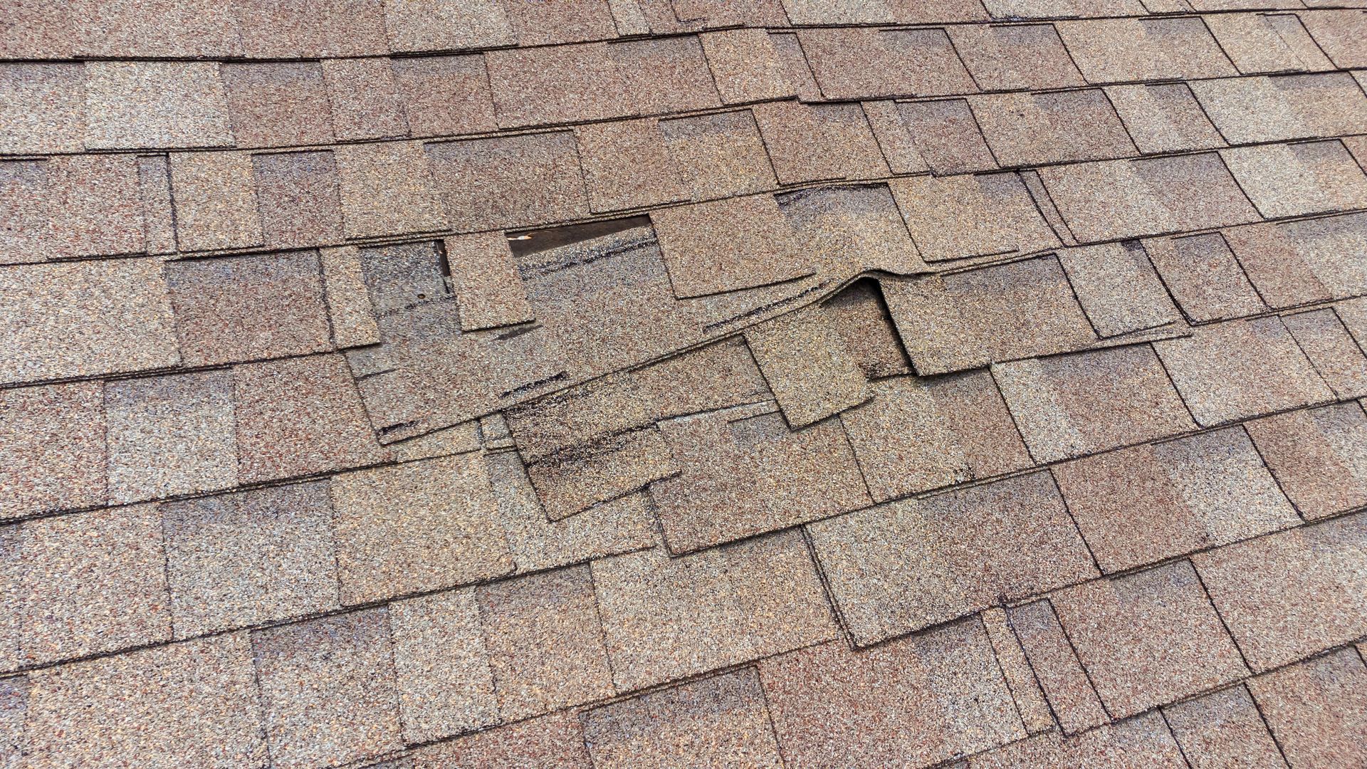 Damaged brown asphalt roof shingles with a section missing and some overlapping.