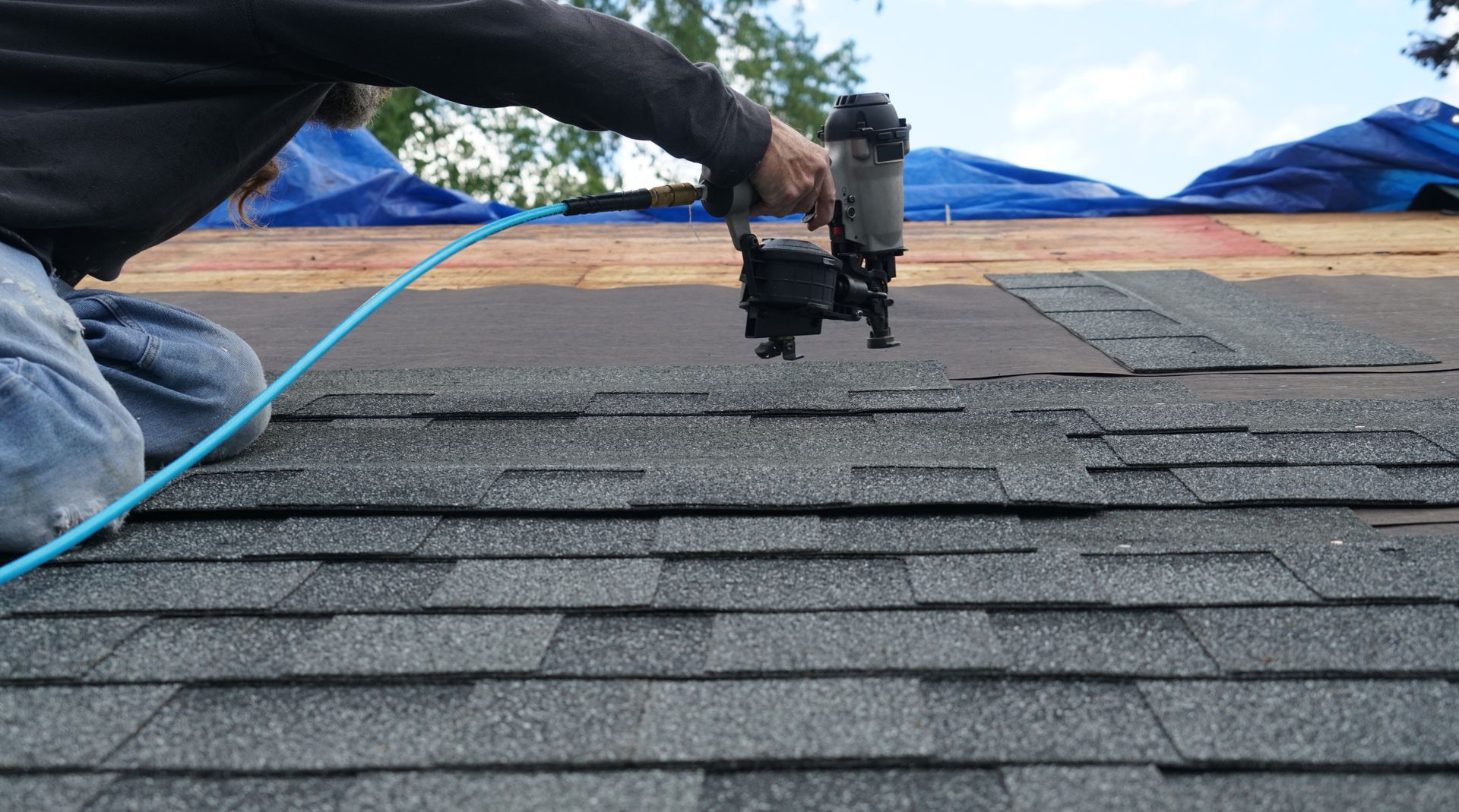 Roofer using a nail gun on an asphalt shingle roof; blue hose connected to the gun.