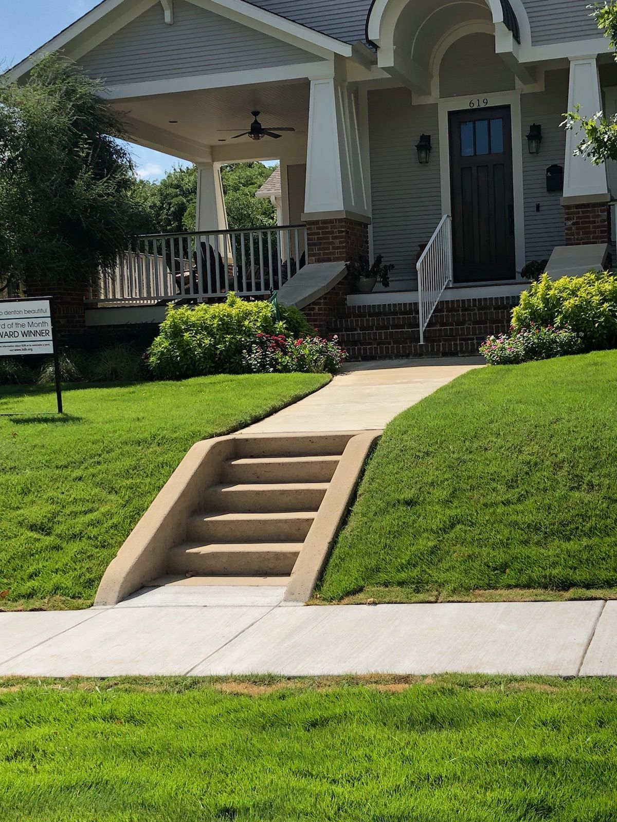 Front yard with a concrete walkway, grass slopes, and stairs leading up to a porch of a house