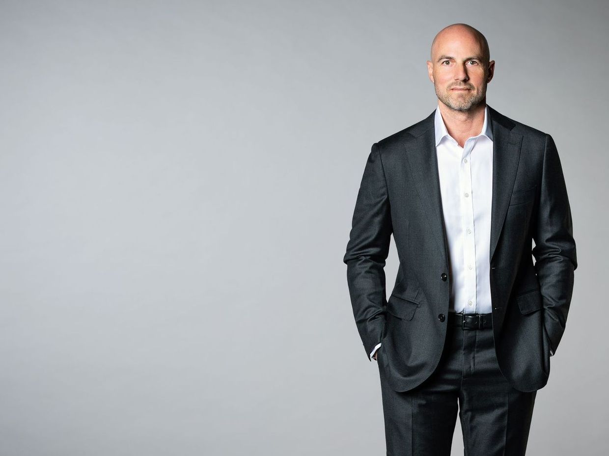 A bald man in a suit and white shirt is standing with his hands in his pockets.