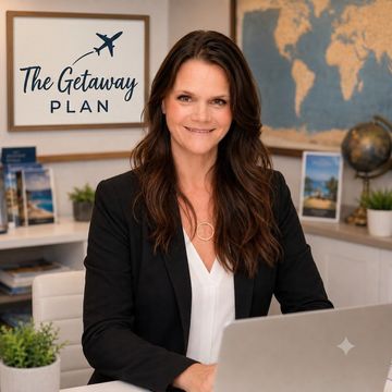 Woman at a desk with a laptop, smiling in an office with a “The Getaway Plan” sign and travel decor.