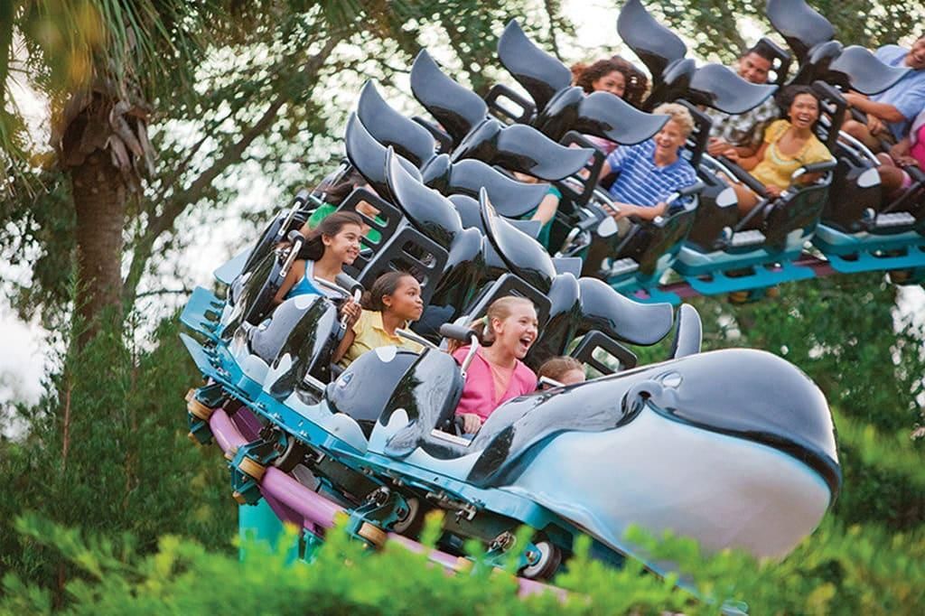 People riding a shark-themed roller coaster through a green park.