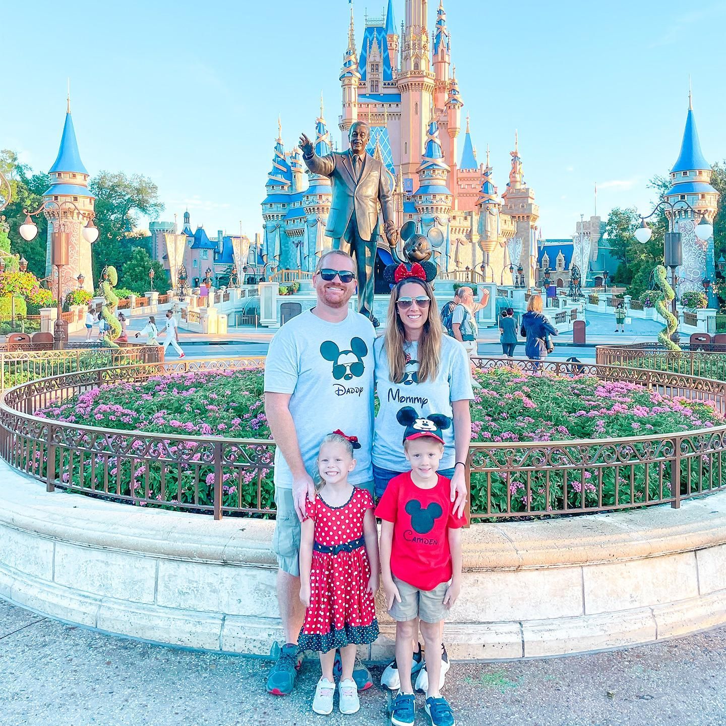 Family posing in front of a colorful fairytale castle at a theme park, with flowerbeds in the foreground.