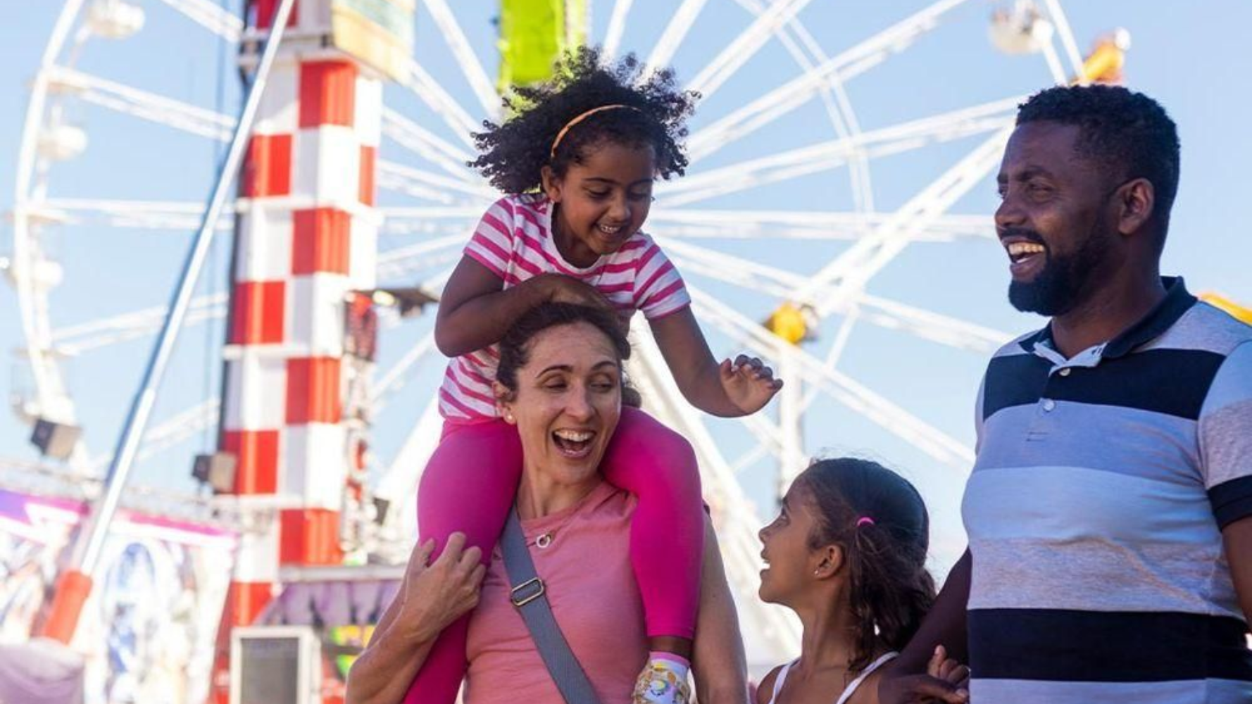 Family smiling at a carnival, with a child on an adult’s shoulders near a Ferris wheel.