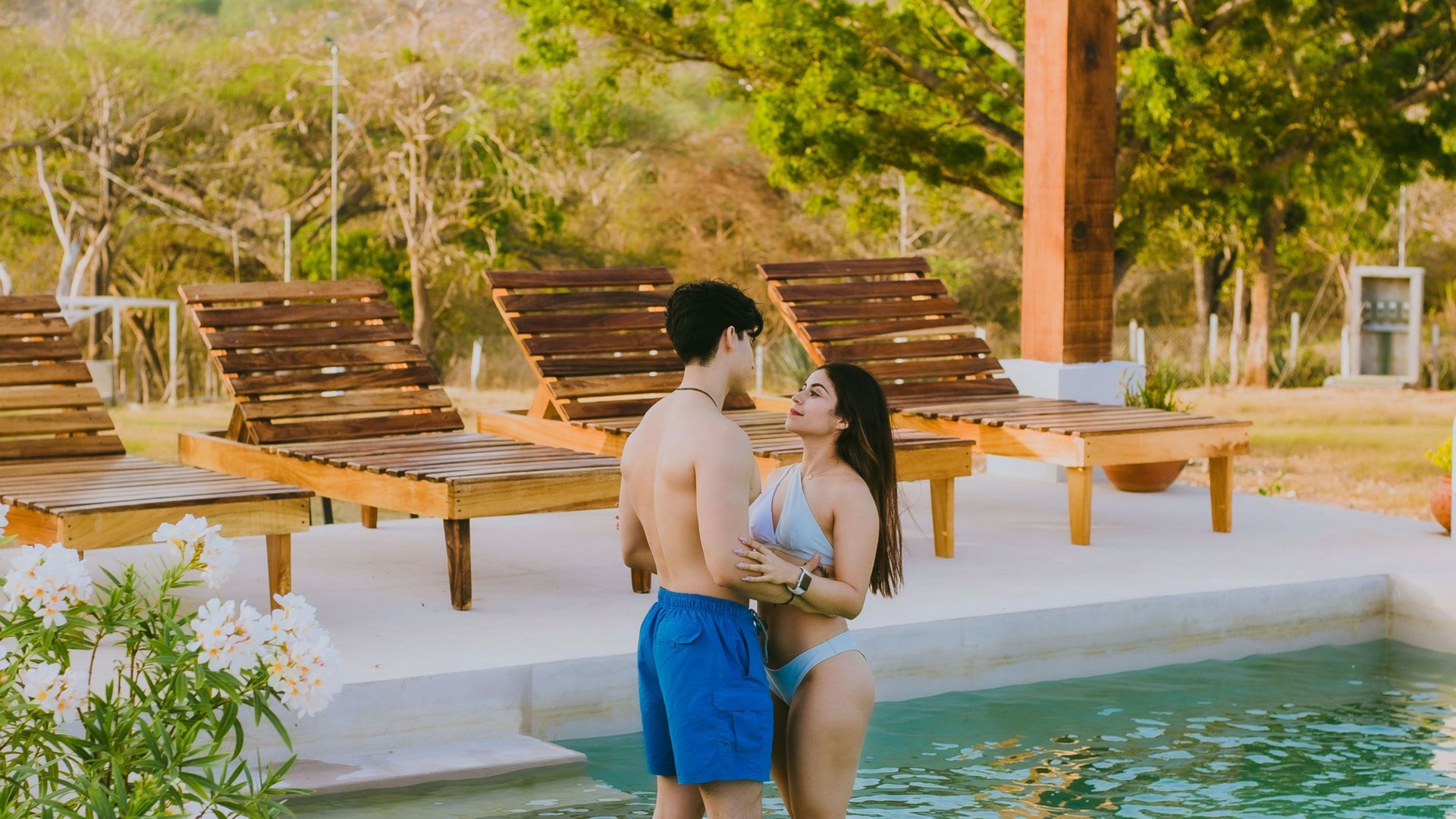 Two people standing in a pool beside wooden lounge chairs at a sunny resort.