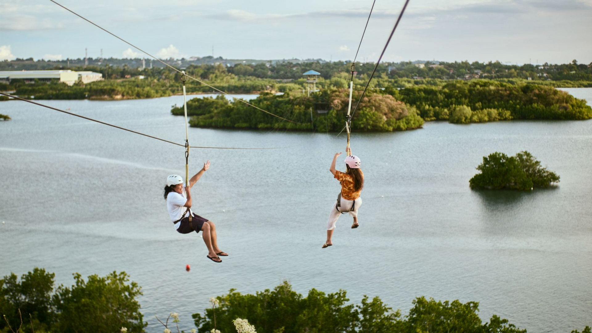 Two people ziplining over a calm lake with small islands and green trees in the background