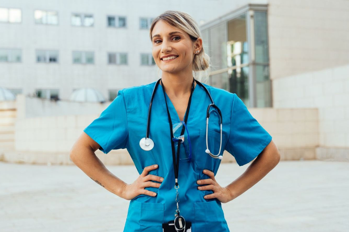 A nurse with a stethoscope around her neck is standing with her hands on her hips in front of a hospital.