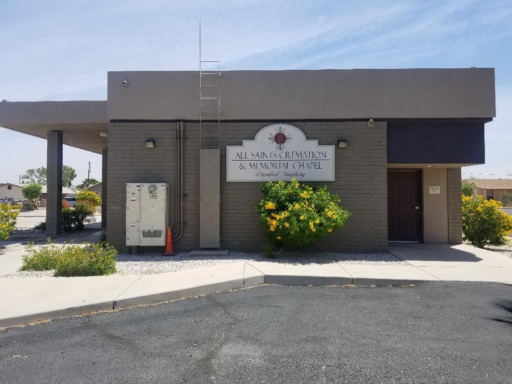 Small gray building with a sign, ladder, flowers, and a parking lot in front.