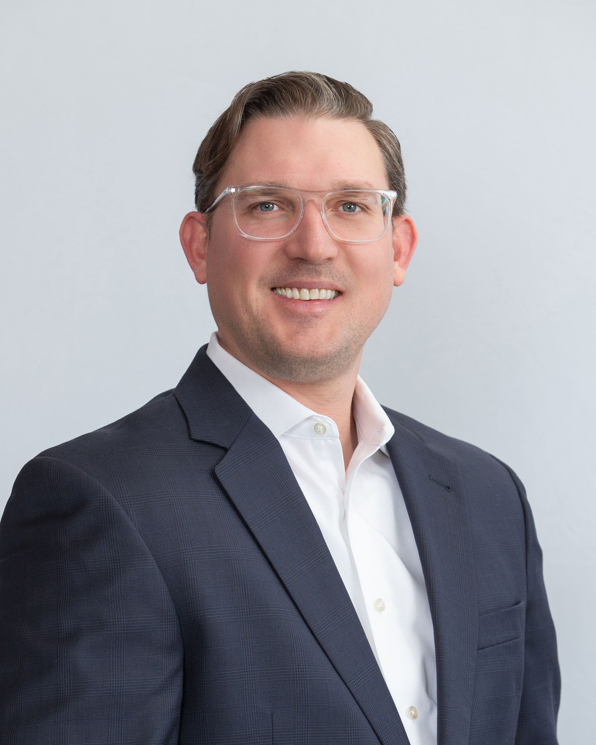 Professional headshot of a smiling person in a dark blazer and white shirt against a light gray background