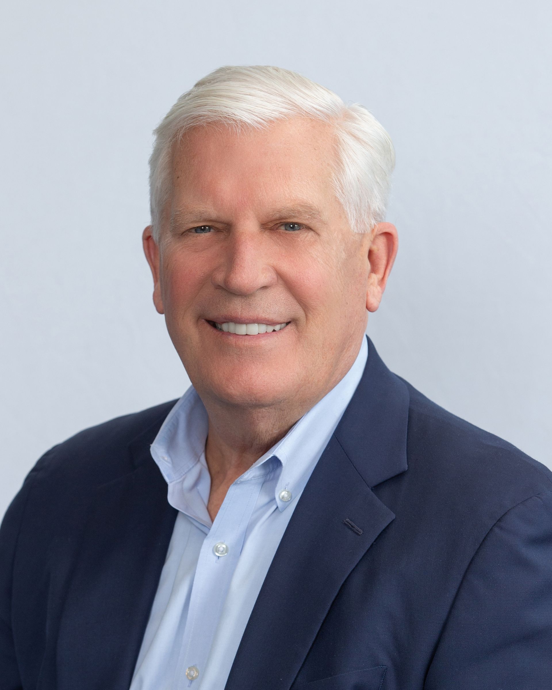 Smiling man in a navy blazer and light blue shirt against a light gray background